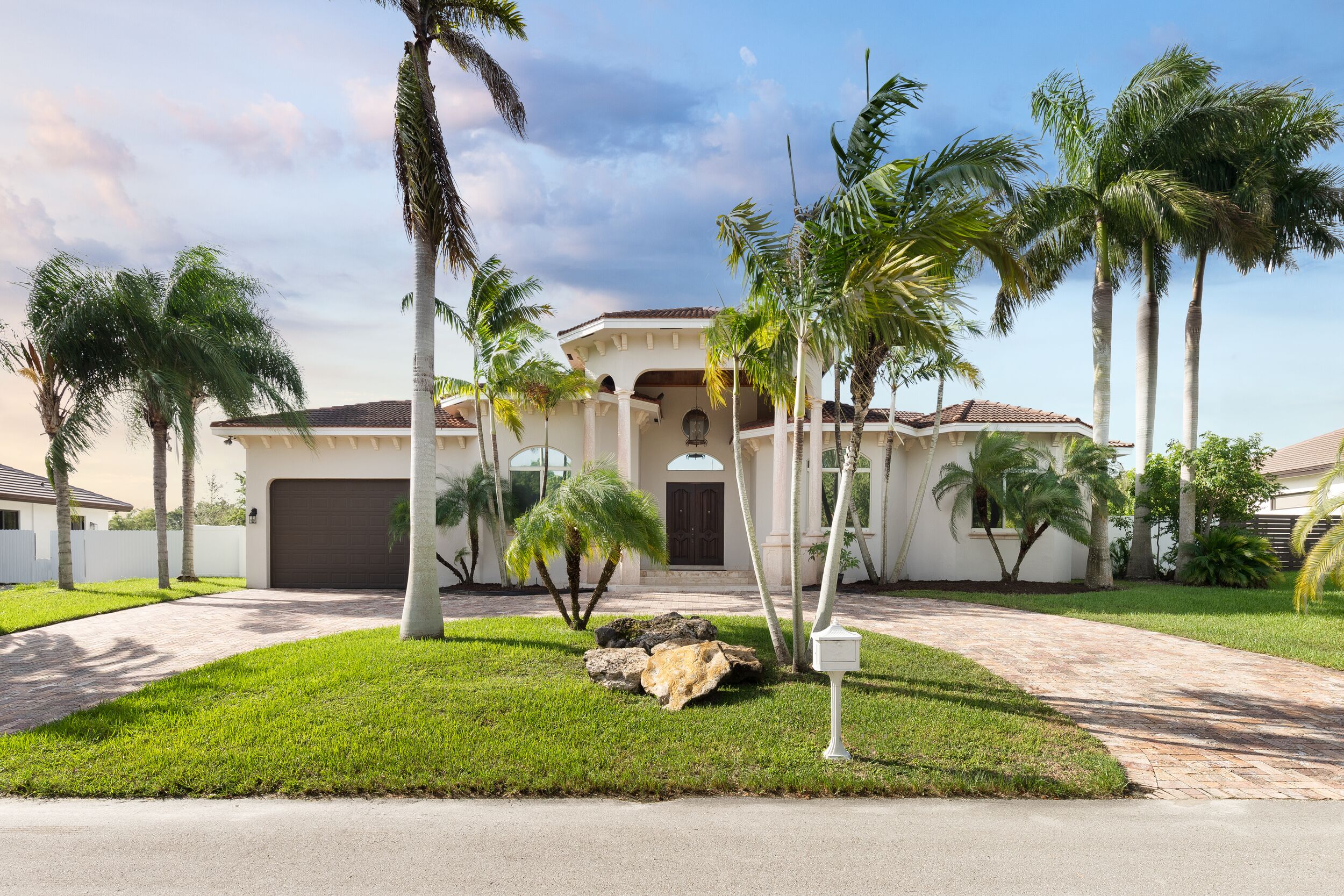 Facade of the entrance to a mansion with wooden doors, garage, palm trees, grass and a blue sky.